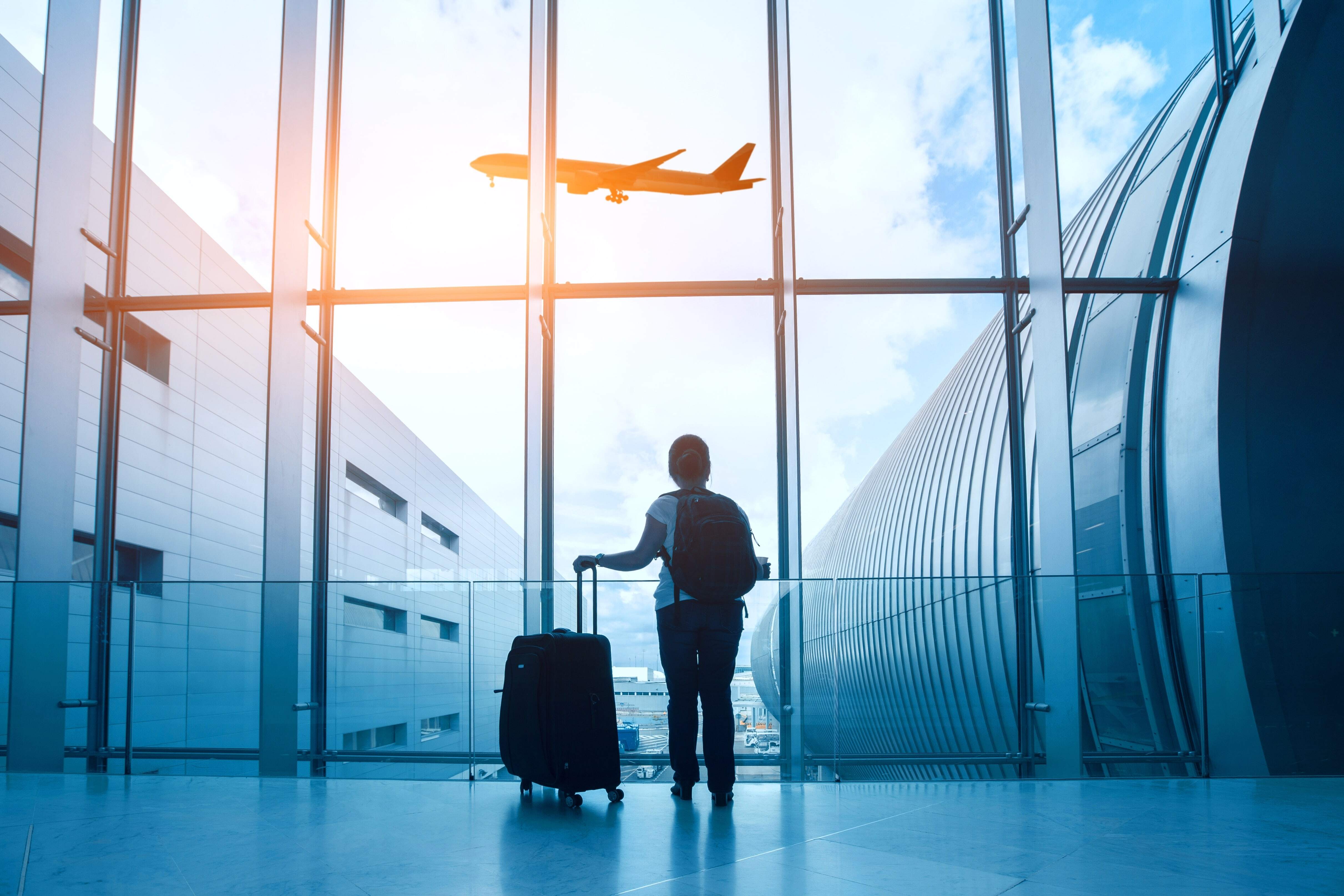 person standing with a suitcase looking out an airport window at an airplane taking off